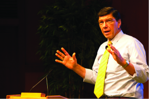 A man with glasses, a white shirt and yellow tie is speaking at a podium.