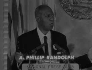 In a black and white image, Philip Randolph stands at a podium wearing a suit and tie, speaking to an audience. The podium is labeled "National Press Club" and he is looking off to the right.