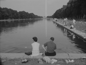 A black and white photo shows a couple sitting with their back towards the camera. Their feet are soaking in the water directly across from the Washington Monument.