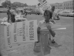 A black and white image shows a woman holding signs reading statements like "Civil Rights" and "Freedom". In the background, other protestors can be seen holding and making other signs.
