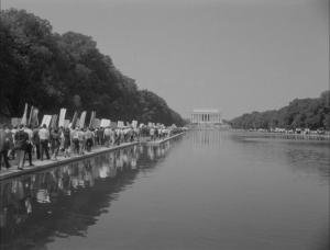 A black and white photo depicts protestors marching towards the Lincoln Memorial. The protestors are to the far left of the image holding signs, and the memorial is visible in the background.