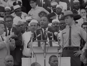 Martin Luther King Jr. stands behind a podium speaking to a crowd at the March on Washington. He is surrounded by protestors standing behind him.