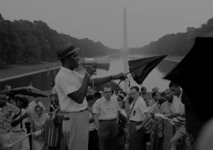 A black and white image shows protestors standing across from the Washington Monument, which is visible in the background of the photo. Protestors gather around a man speaking into a megaphone and gesturing with an umbrella. He is wearing a fedora, a white shirt, and grey pants, and he is staring off to the right.