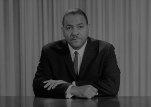 A black and white image shows Carl T. Rowan sitting behind a desk, in front of curtains. He is wearing a suit and tie and staring directly into the camera.
