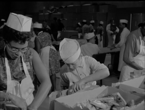 A woman and child stand next to each other bagging sandwiches. They are wearing white hats and aprons and looking down at their work.