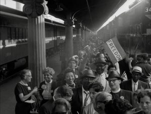 A black and white image shows a crowd gathered in a train station. In the midst of the crowd, a person is holding a flag that reads "Freedom".
