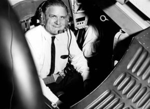 A black and white image shows James Webb in a cockpit, wearing a tie, white shirt, and headset.