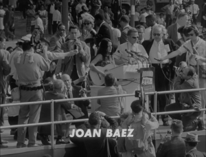 A black and white image shows Joan Baez at a podium, holding a guitar. She is surrounded by a crowd.