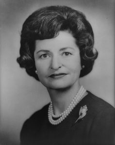 A black and white image shows Lady Bird Johnson wearing pearls and staring into the camera.