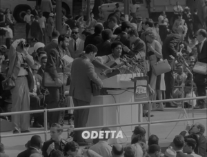 A black and white image shows Odetta behind a podium. She is holding a guitar and surrounded by a crowd.