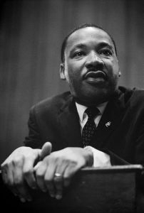 A black and white image of Martin Luther King Jr. shows him wearing a suit and tie, and staring off to the right of the image.