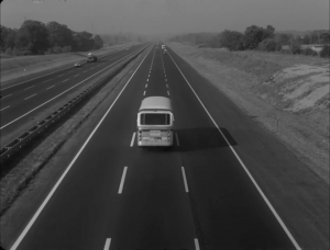 A black and white image shows a bus centered in the middle of a freeway.