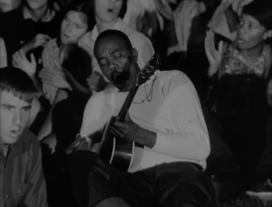 A black and white image shows a man playing guitar in the midst of a crowd.