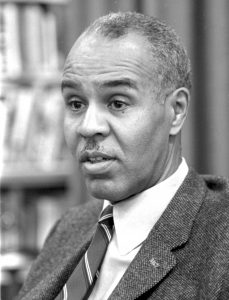 A black and white image shows Roy Wilkins wearing a suit and tie and looking off to the left of the image, in an interview.