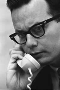 A black and white image shows Bill Moyers with black glasses and speaking on a white phone.