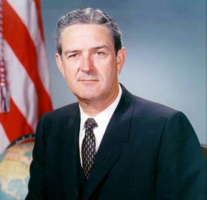 The image shows John Connally posing in front of an American flag and a globe. He is wearing a suit and tie.