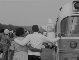 A couple stands arm-in-arm facing the capitol building. The man has his hand placed on the frame of a bus and other protestors can be seen in the distance.