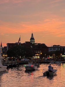 Annapolis waterfront at sunset, showing the dome of the Maryland capitol rising amid waterfront buildings, with boats in the foreground. Above the city, the sky glows orange.