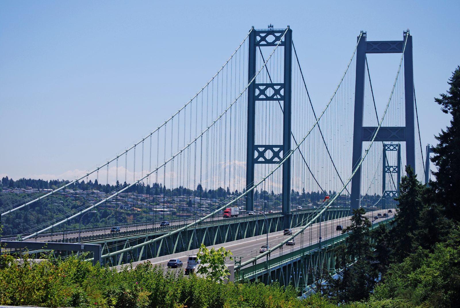 Two twin suspension bridges, known as the Tacoma Narrows Bridge, on a sunny day. Greenery is seen in the foreground, and cars drive over each bridge in the correct direction.