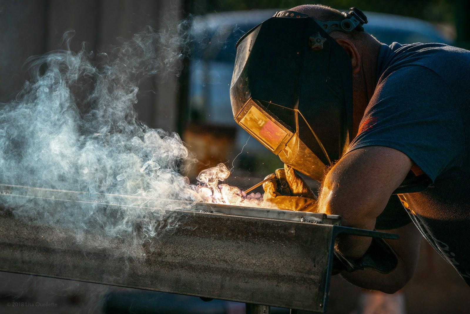 A man performs SMAW welding in a short sleeved T-shirt, gloves, and a welding hood.