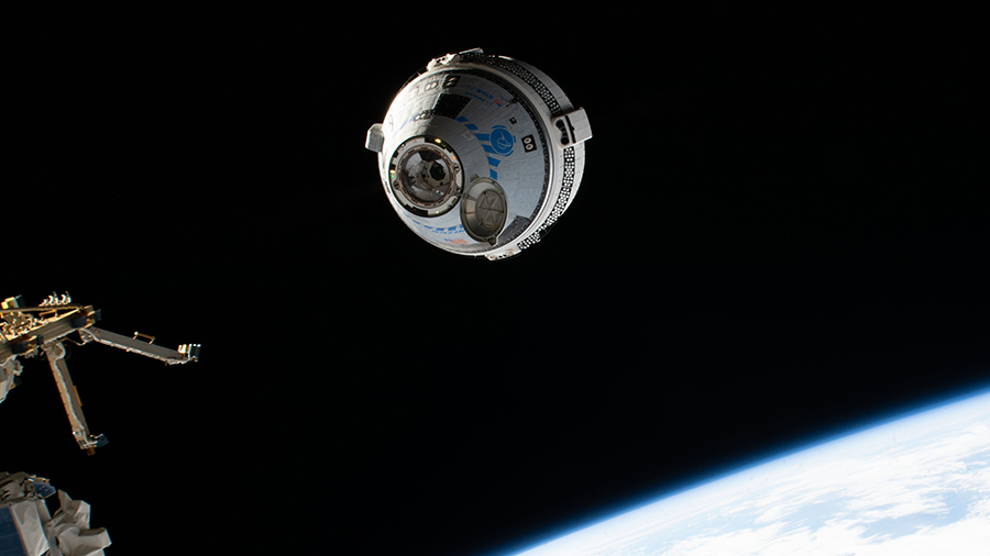 A space capsule in space. Earth is pictured on the lower right. The capsule is cylindrical with a cone-shaped front. To the left, and slightly out of frame, is the International Space Station prepping to accept the crew capsule.