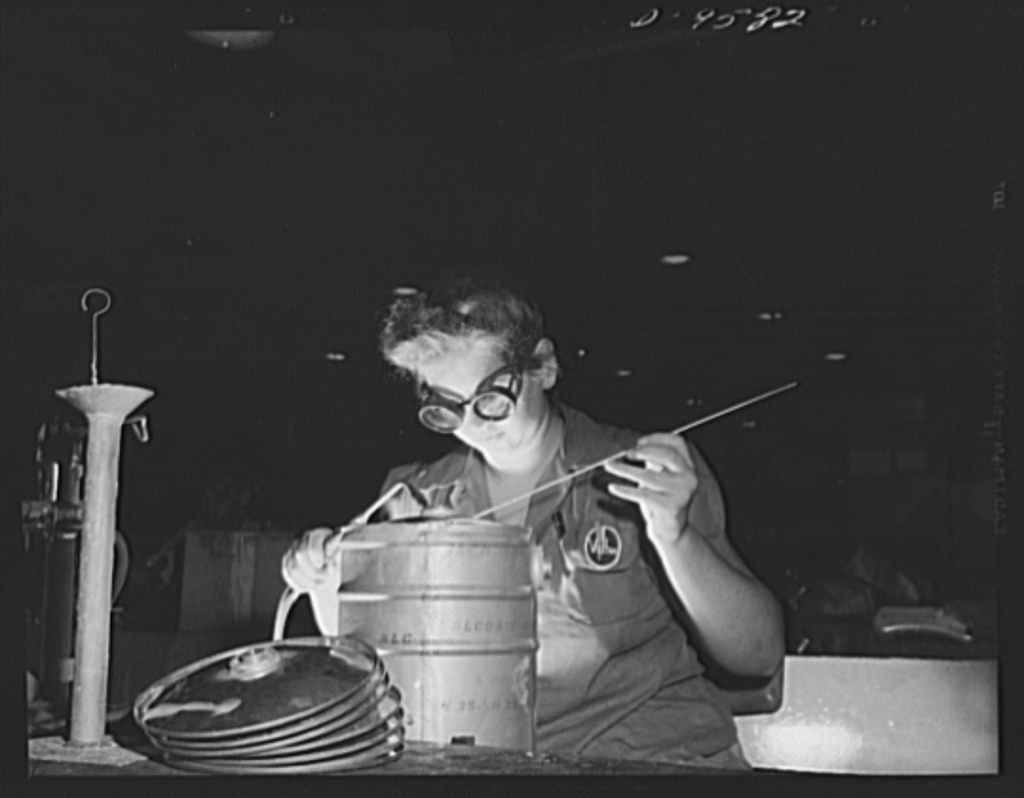 A World War Two era woman, wearing a pair of protective glasses, and sitting comfortably at a work table holds a torch in her right hand and an electrode in her left hand as she focuses on welding an aluminum canister. This is an example of aluminum oxy-acetylene welding.