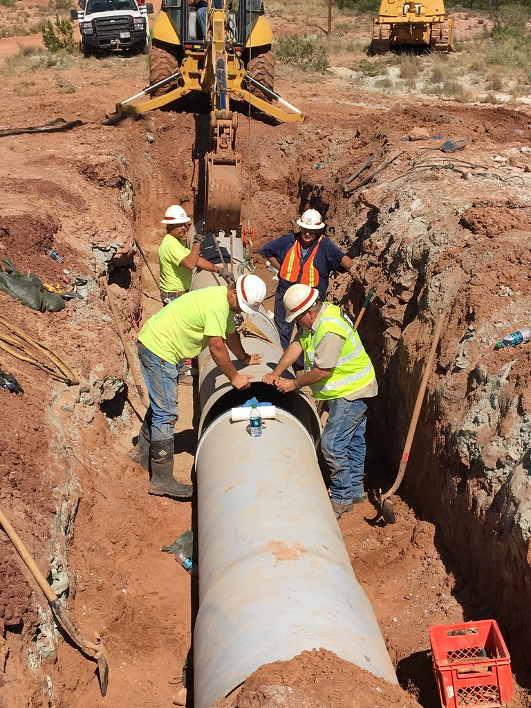 Three workers in a trench working to align large pipe sections for welding.