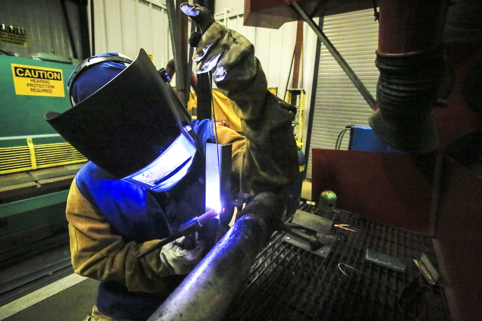 A person wearing a protective welding jacket and welding hood, welding a practice piece vertically using GTAW