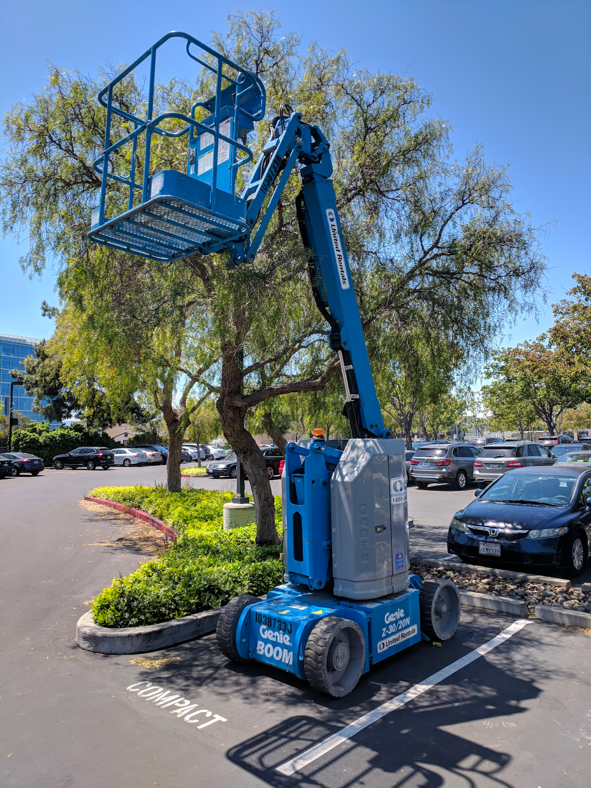 A small lift with an articulating arm is parked outside in a parking lot next to a car.