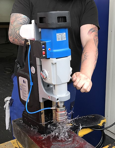 A person holding a magnetic drill on a piece of metal while operating the drill. The drill has a vertically mounted motor that slides up and down on a track, and is attached to a large vertical base with a magnet on the bottom. The spindle is mounted in line with the motor so that it is pointed downward. The operator is turning a handle on the back side of the machine which is forcing the drill down into the work piece.