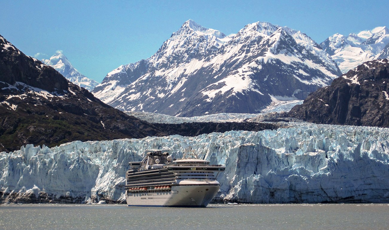 A cruise ship as it sails in front of a snowy glacier with a dramatic mountain in the background.