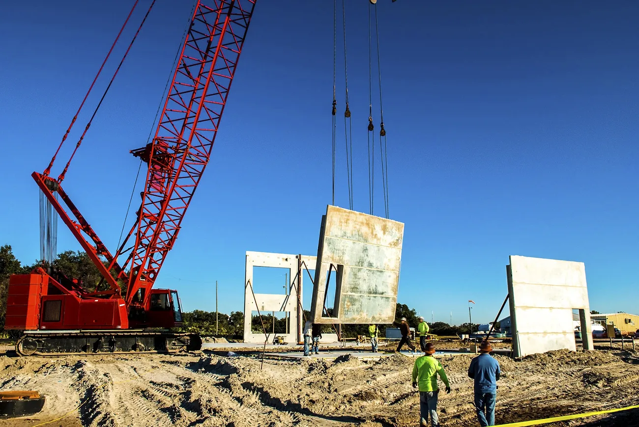 A lattice boom crane mounted on crawler treads lifts precast tilt-up concrete panels into place on a construction site.
