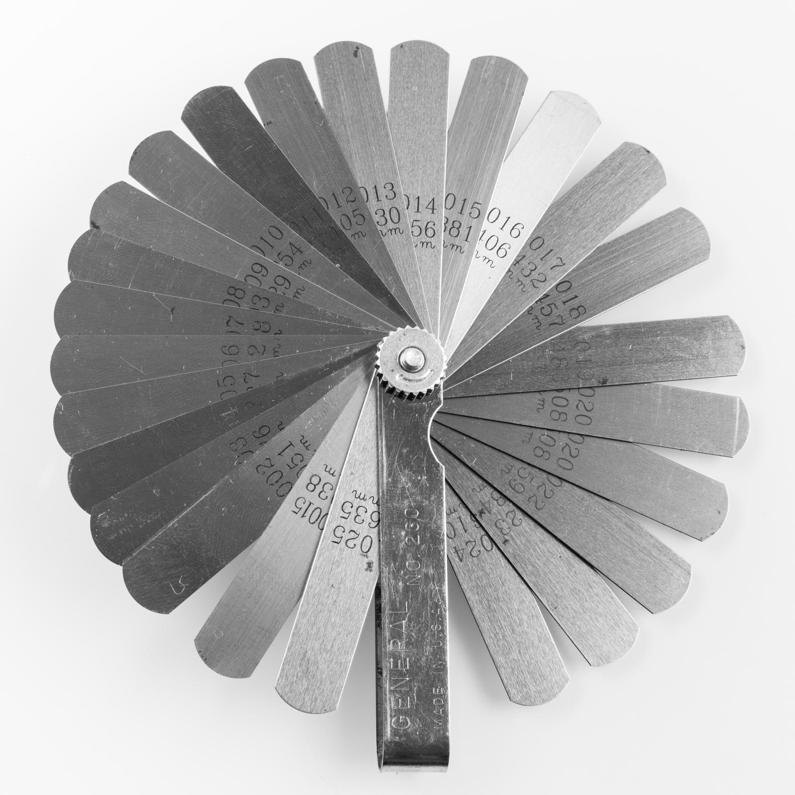 A set of feeler gauges fanned out. Each feeler gauge is a thin strip of metal with a measurement printed on it. The gauges are held together with a nut at the base so that each gauge can be inserted in a gap but stay attached to the entire set.