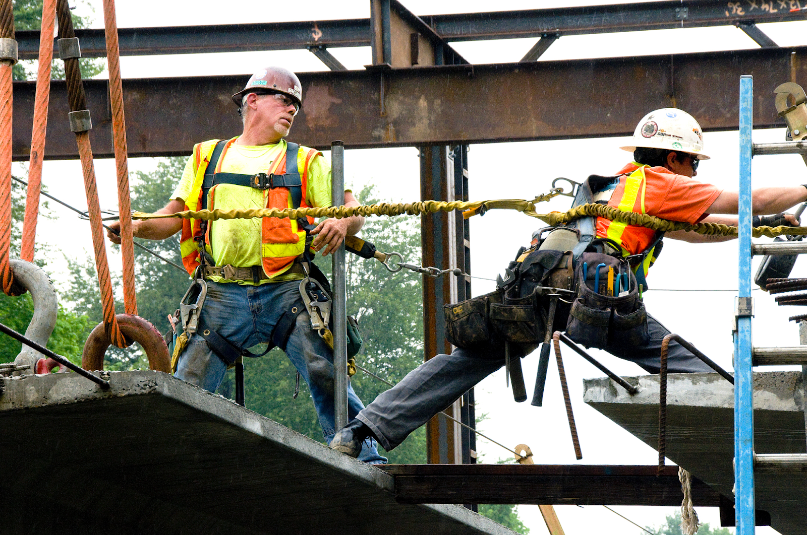 Two individuals work on a construction site. One person reaches across a platform to secure his lanyard to a second position.