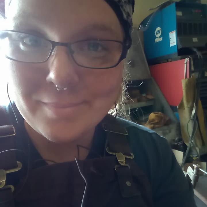 Jessica Y, smiling and wearing welding gear, glasses, and a nose ring in a shop environment.