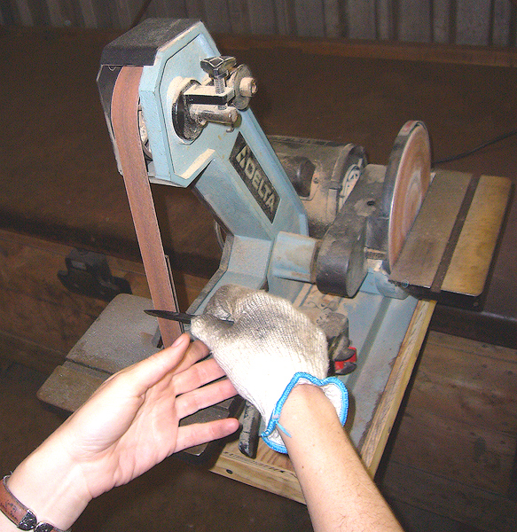 A small tabletop belt sander. A person wearing a glove holds a piece of metal against an abrasive belt that rapidly rotates as it moves around two drums.