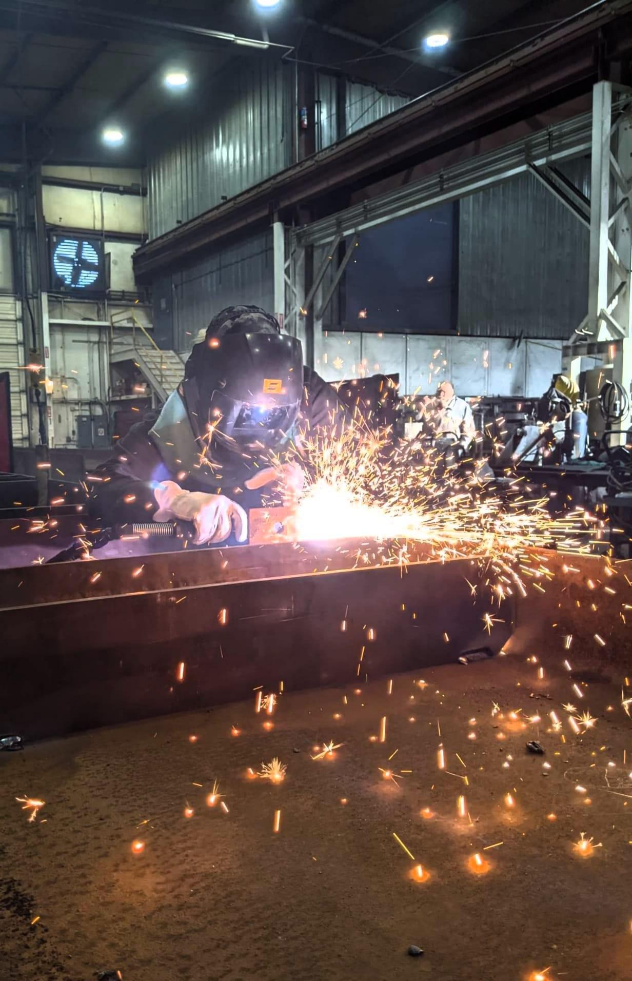 Jessica Y wearing a welding hood, jacket, and leather gloves uses a plasma cutter to trim a part on a box shaped weldment.