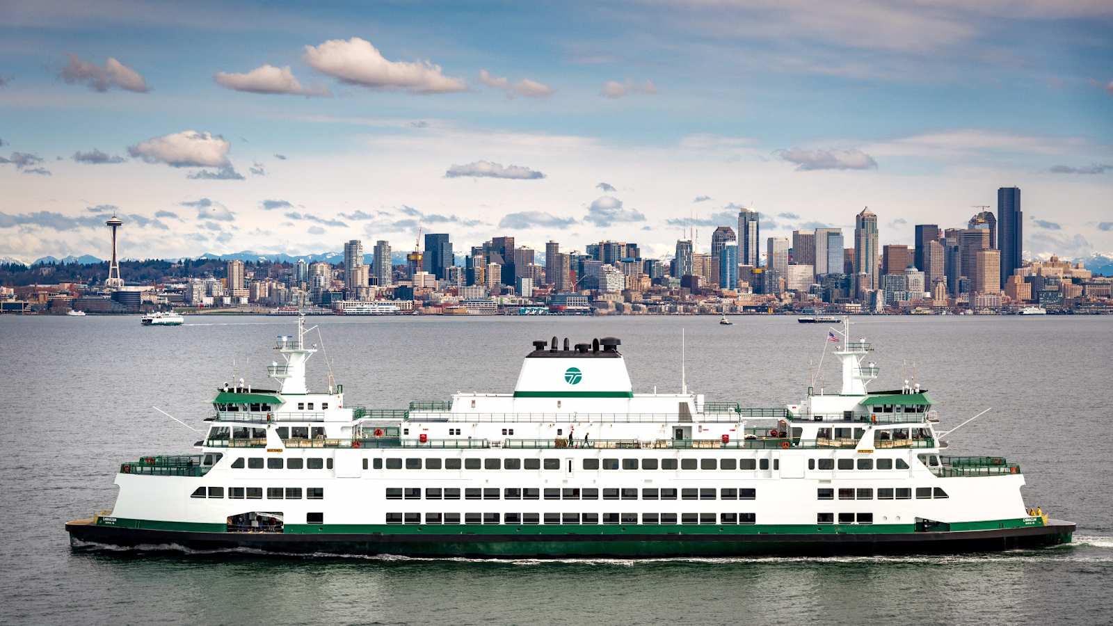 Washington State Ferry is in the foreground is the. The ferry is white, with green detailing at the waterline and on railings. In the background is the city of Seattle skyline.