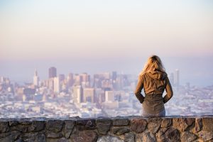 In the foreground, a person sits on a stone wall, gazing at a city in the background.